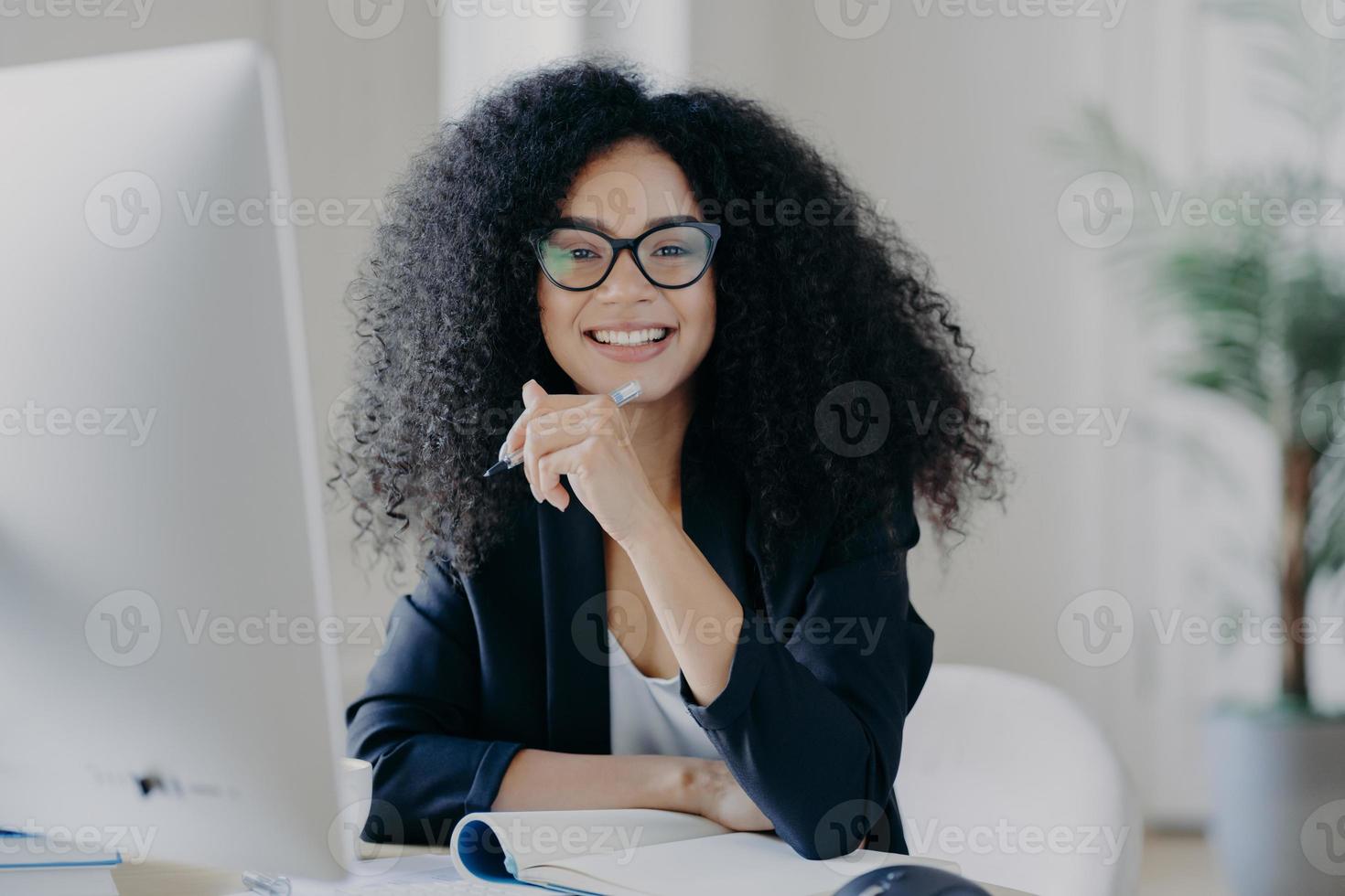 positive internationale studentin mit krausem haar, trägt durchsichtige brille, hält stift in der hand, macht buchhaltung, sitzt vor großem computerbildschirm, gekleidet in schwarzes elegantes outfit. foto