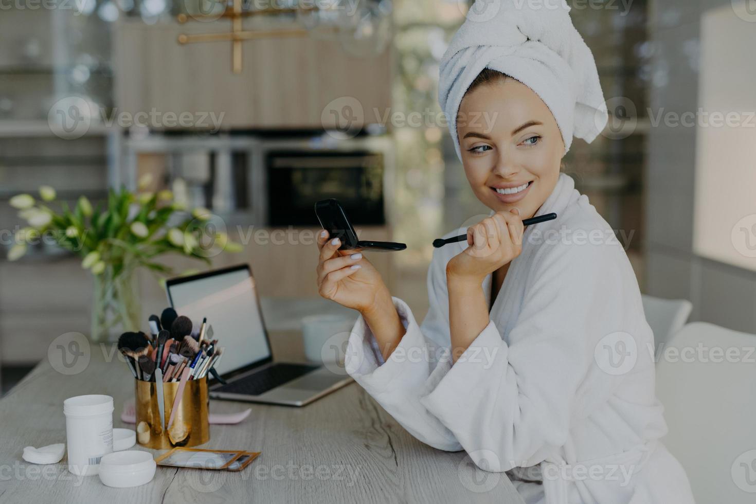 innenaufnahme einer lächelnden frau schminkt sich nach dem duschen, hält kosmetikpinsel und spiegel trägt bademantel und eingewickeltes handtuch auf dem kopf. frauen, die wellness- und schönheitsbehandlungskonzept pflegen foto