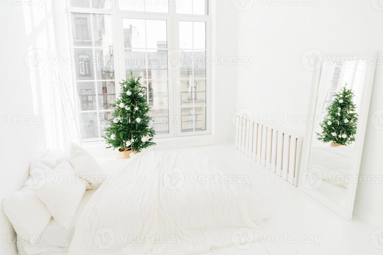 komfort-, zuhause- und heiligabendkonzept. Schlafzimmer mit weißem, weichem Bett, Spiegel, großem Fenster für Lichteinfall, geschmücktem Neujahrsbaum und Leiter. Feiertagsdekoration. foto