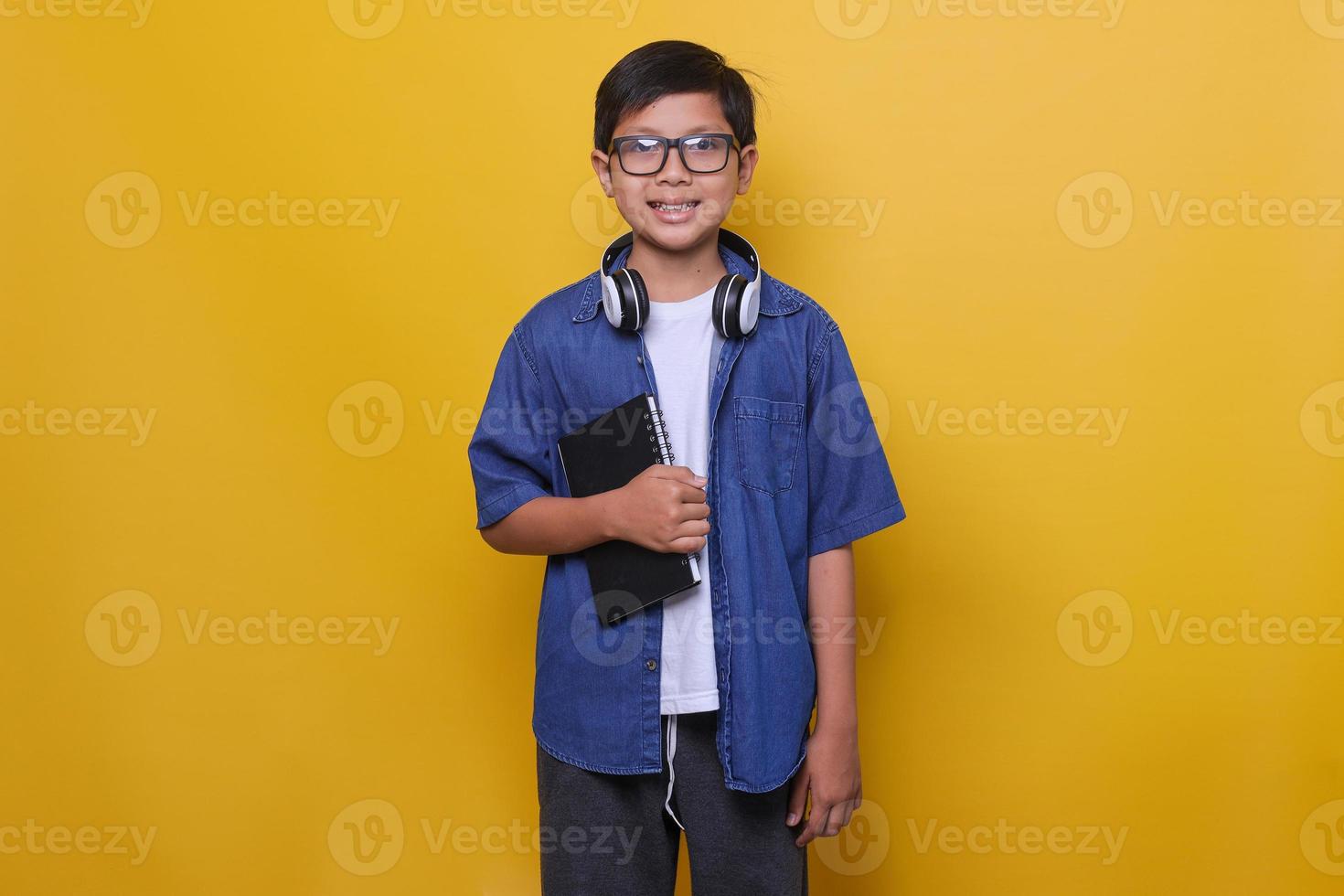 smart kid in lässiger kleidung, kopfhörer und brille steht und hält buch, lächelt in die kamera auf gelbem hintergrund. foto