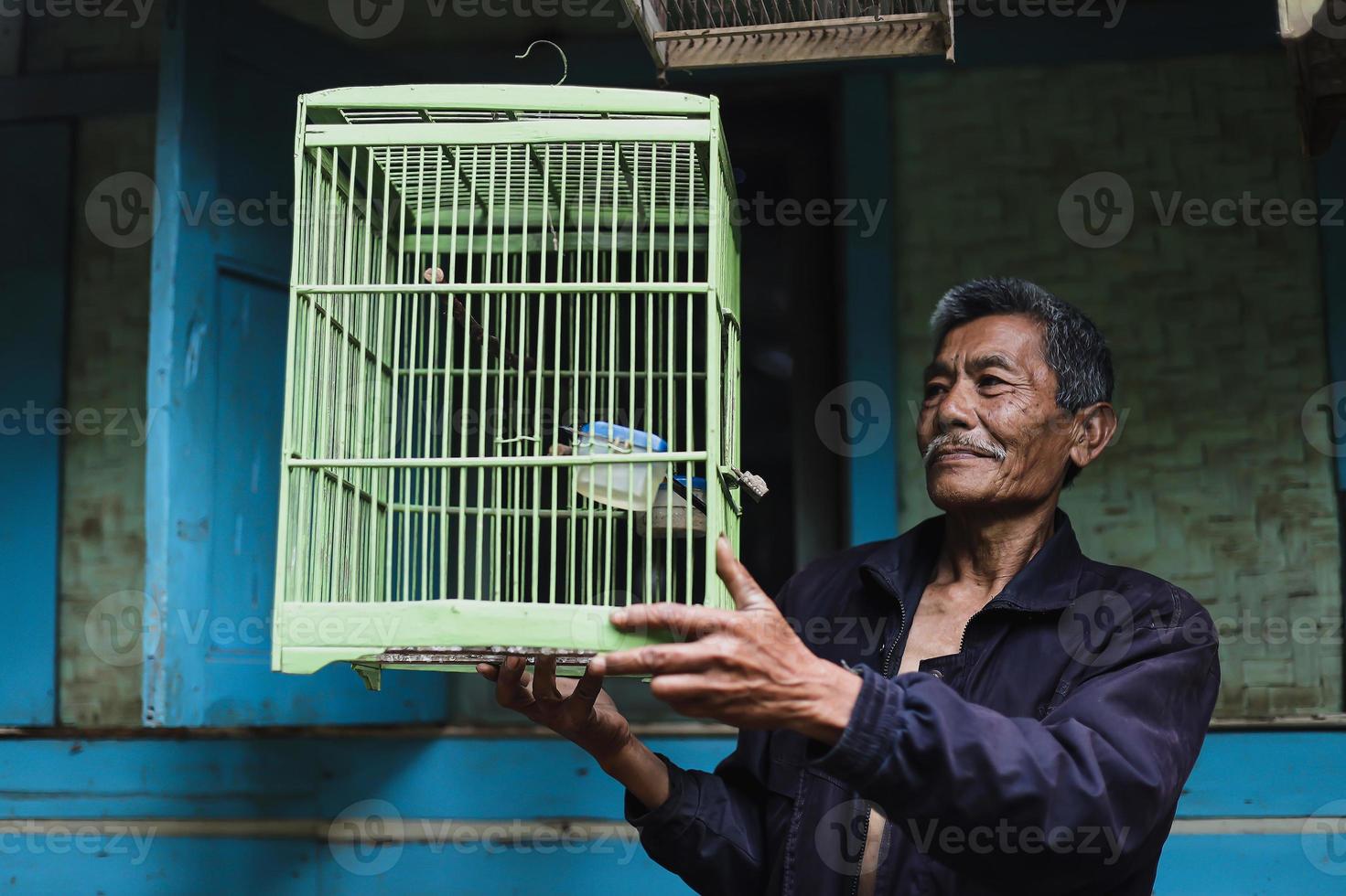 asiatischer älterer mann, der grünen vogelkäfig außerhalb des traditionellen holzhauses hält. foto