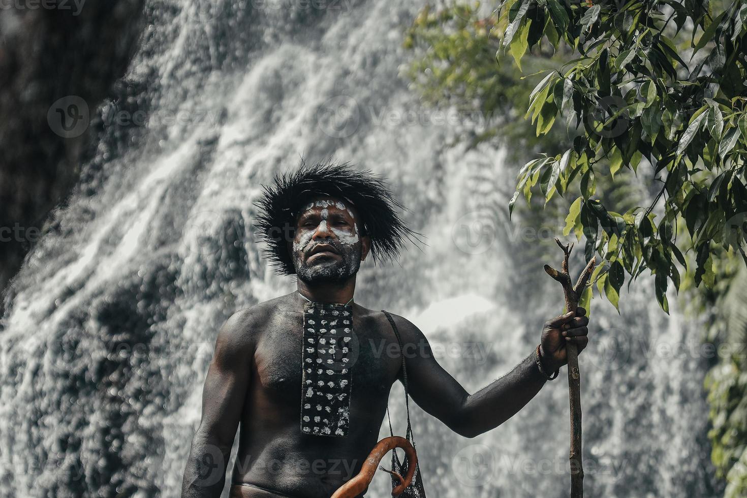 papua-mann des dani-stammes, der gegen verschwommenen wasserfall im grünen wald steht foto
