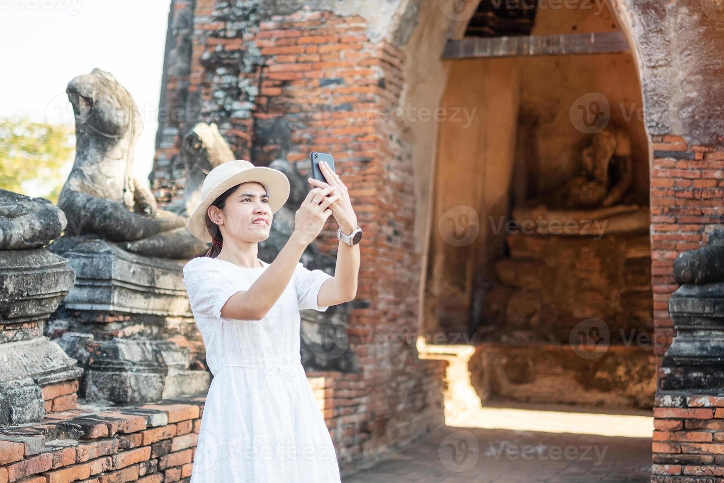 glückliche touristische frau im weißen kleid, das foto mit mobilem smartphone macht, während des besuchs im wat chaiwatthanaram-tempel im historischen park ayutthaya, sommer-, solo-, asien- und thailand-reisekonzept