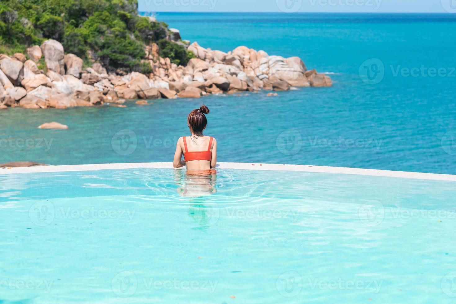 glückliche Frau in orangefarbenem Badeanzug, die im Infinity-Pool im Luxushotel am Meer schwimmt. junge frau genießt im tropischen resort. entspannendes, sommer-, reise-, urlaubs-, urlaubs- und wochenendkonzept foto