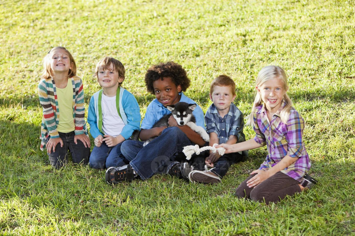 gemischtrassige Kinder, die auf Gras mit Huskywelpen sitzen foto