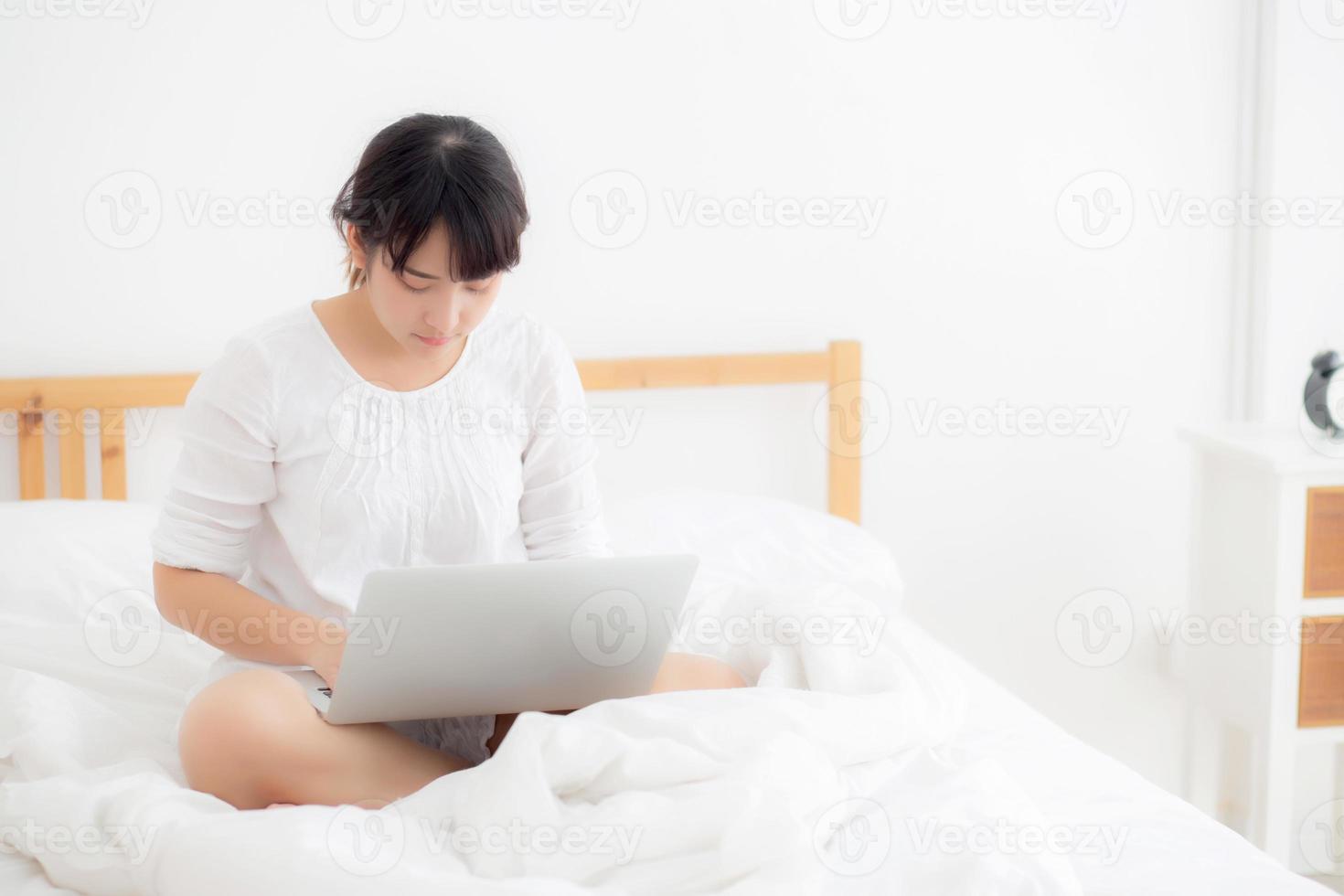 porträt einer schönen asiatischen jungen frau, die auf dem bett sitzt und einen laptop im schlafzimmer für die freizeit und entspannung verwendet, freiberuflich mit mädchen, die notizbuch, kommunikations- und lebensstilkonzept arbeiten. foto