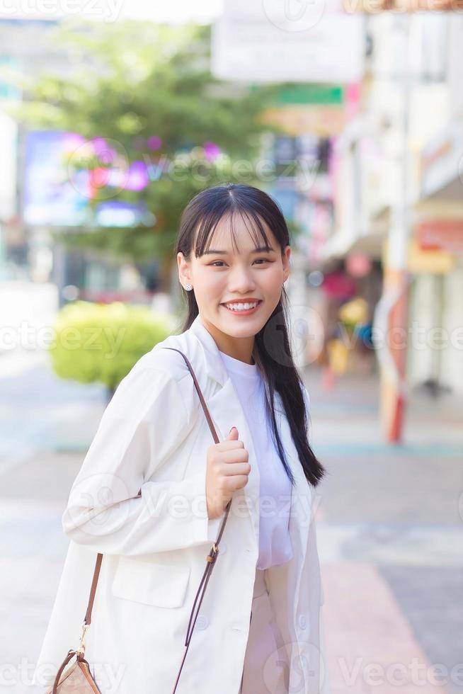 selbstbewusste junge asiatin, die ein weißes hemd und eine umhängetasche trägt, lächelt glücklich, während sie zur arbeit im büro in der stadt geht. foto