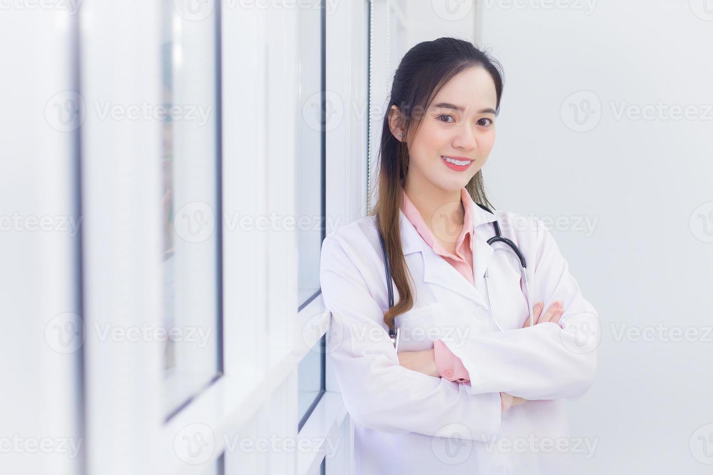 professionelle asiatische ärztin mit schwarzen langen haaren trägt einen weißen laborkittel und ein stethoskop. sie steht und verschränkt die arme im büro im krankenhaus. foto