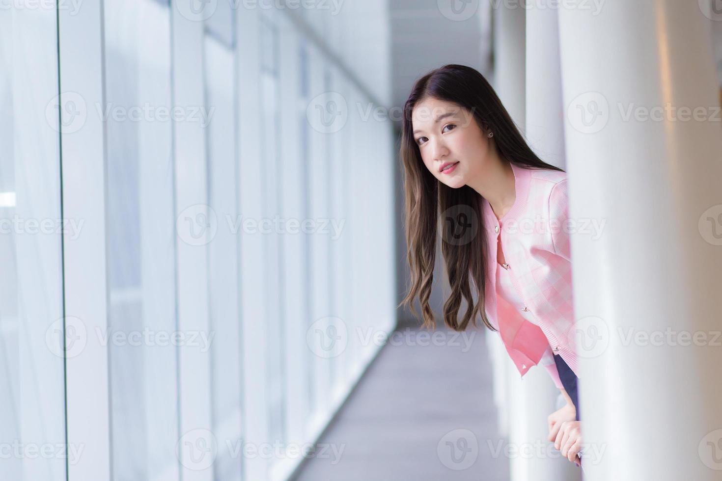 Schönes asiatisches Teenager-Mädchen im Teenageralter trägt ein weißes mit rosafarbenem Hemd und lächelt fröhlich, während sie an einem schönen Tag in der Nähe des verglasten Fensters steht. foto