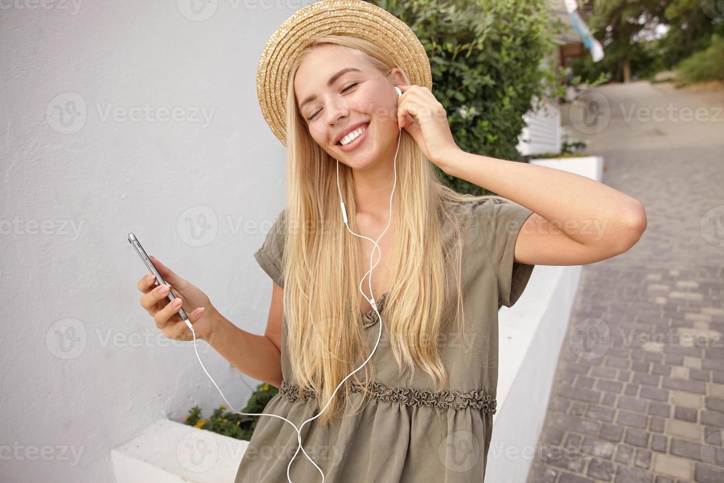 Outdoor-Nahaufnahme einer attraktiven glücklichen Frau, die Musik mit Kopfhörern hört, während sie durch die grüne Straße geht und Musiktitel genießt foto