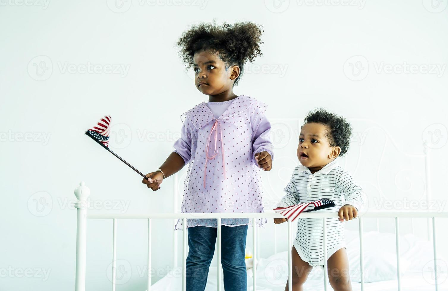 glücklicher süßer aktiver kleiner afroamerikanischer kinderjunge und mädchen, die amerikanische flagge auf dem bett halten foto