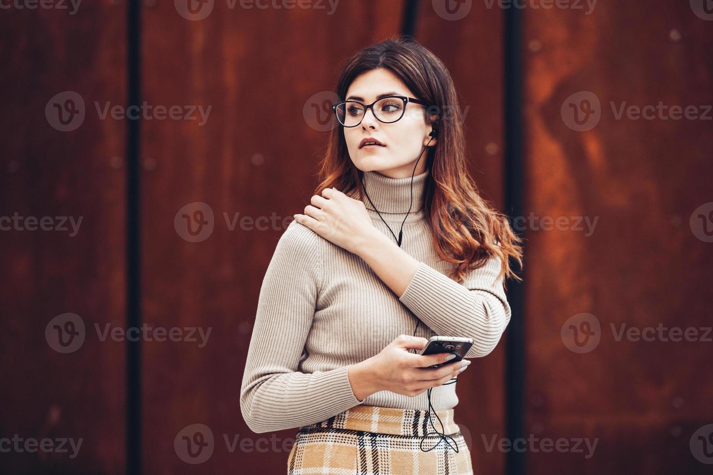 Bild der schönen stilvollen Frau, die Handy hält. junges europäisches mädchen, das auf der straße steht und handy benutzt. frau, die musik mit dem telefon hört und spaß hat. mobiles internetkonzept. foto