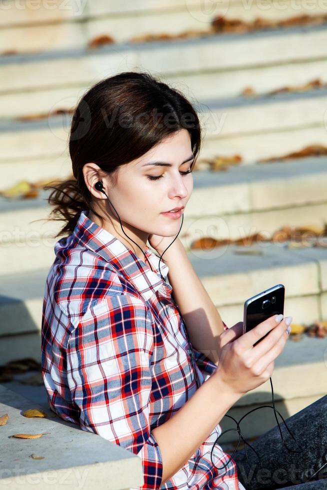 Bild der schönen stilvollen Frau, die Handy hält. junges europäisches mädchen, das auf der straße steht und handy benutzt. frau, die musik mit dem telefon hört und spaß hat. mobiles internetkonzept. foto