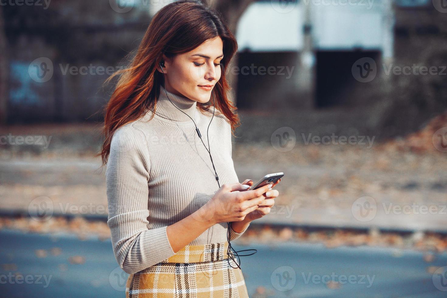 Bild der schönen stilvollen Frau, die Handy hält. junges europäisches mädchen, das auf der straße steht und handy benutzt. frau, die musik mit dem telefon hört und spaß hat. mobiles internetkonzept. foto