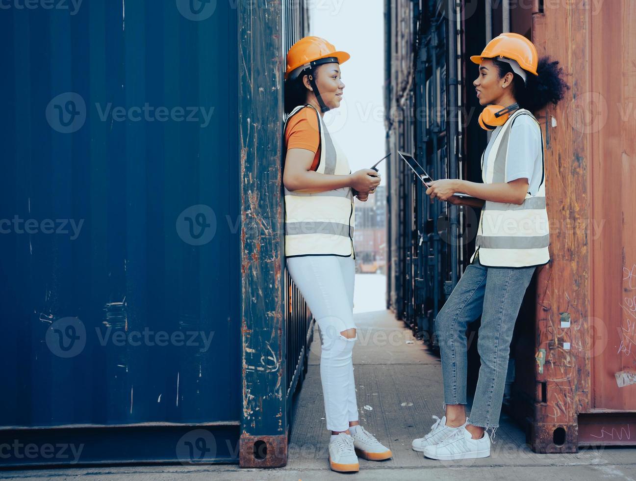 zwei Vorarbeiterinnen am Containerplatz. gemischtrassige frauen, wirtschaftsingenieure stehen mit laptop und walkie-talking-lächelnd in einer containerbox. Internationales Logistik-Frachtterminal. foto