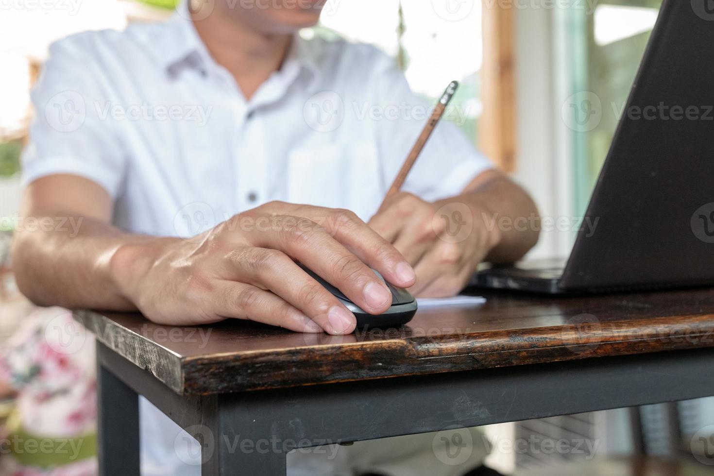 asiatische braune Haut, die eine drahtlose Maus auf dem Vintage-Holztisch hält und mit der linken Hand Bleistift schreibt. foto