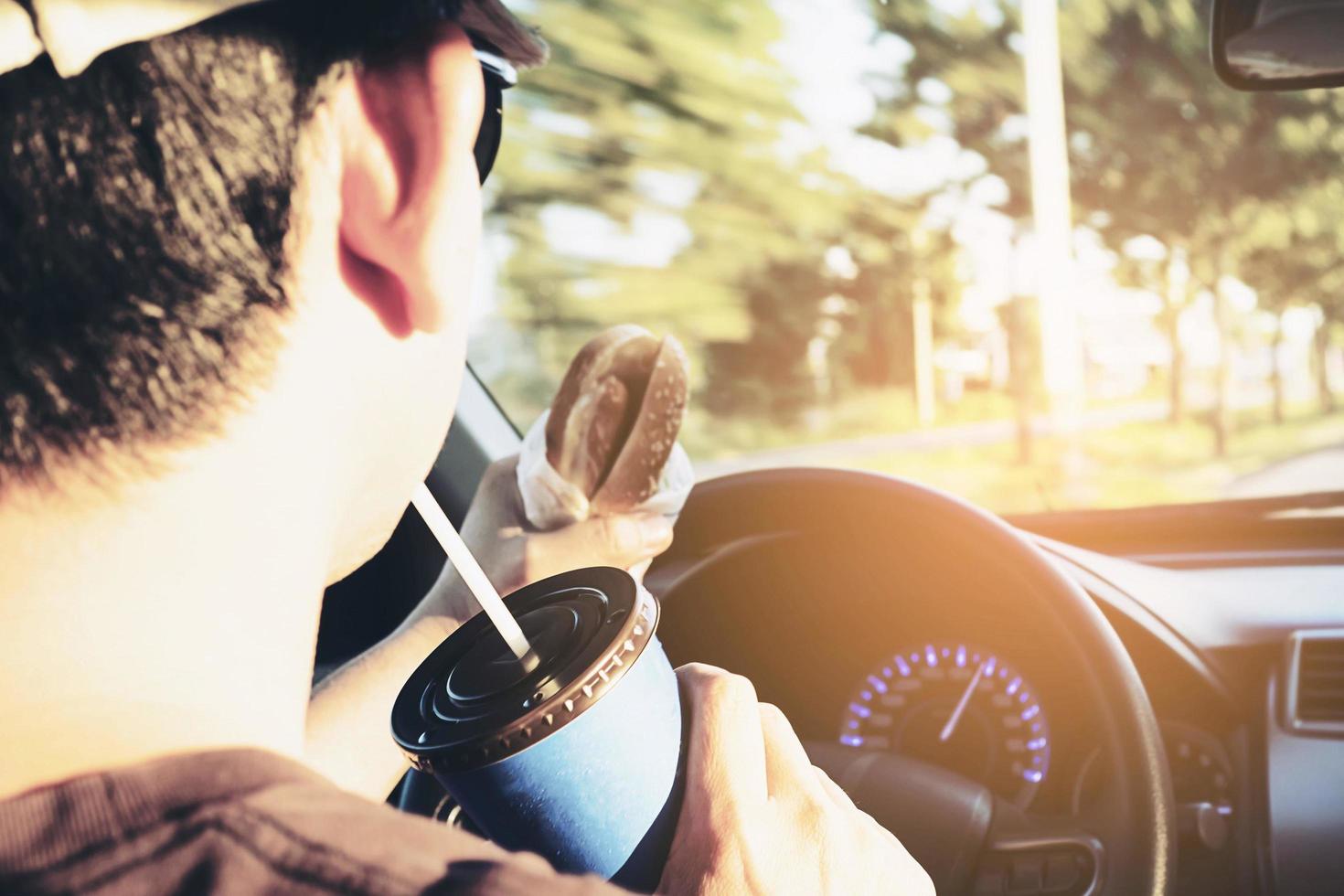 Mann isst gefährlich Hotdog und kaltes Getränk beim Autofahren foto