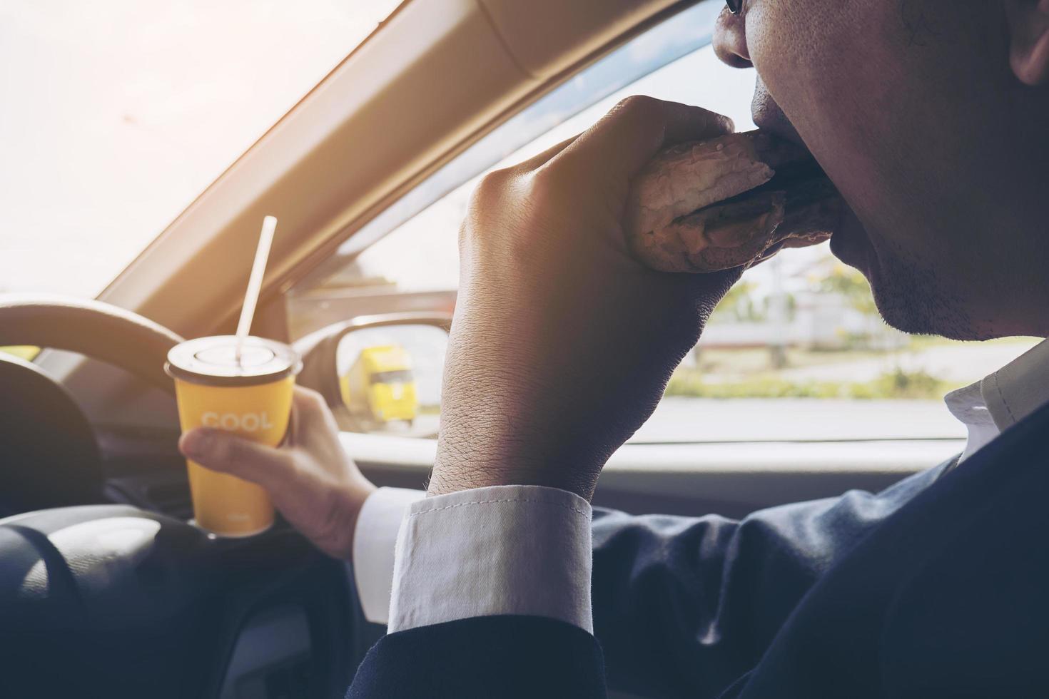 Mann, der Auto fährt, während er eine Tasse kalten Kaffee hält und Hamburger isst foto