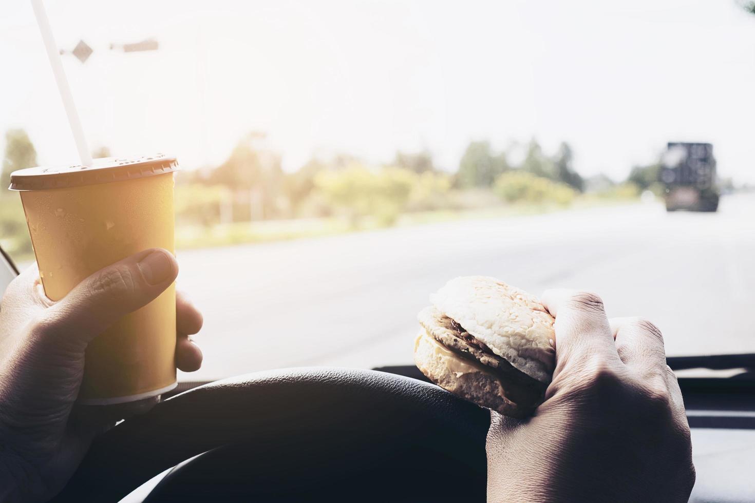 Mann, der Auto fährt, während er eine Tasse kalten Kaffee hält und Hamburger isst foto