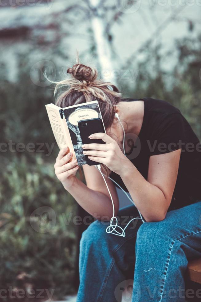 Foto eines fröhlich lächelnden, süßen jungen Studentenmädchens, das im Freien eine Sonnenbrille trägt und mit dem Handy plaudert, um Musik mit Kopfhörern zu hören.