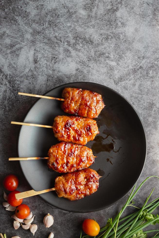 grillen Sie aufgespießtes Milchschwein in einem schwarzen Gericht mit Tomaten mit Knoblauch und Frühlingszwiebeln auf grauem Betonhintergrund. thailändisches straßenessen.draufsicht und kopierraum foto