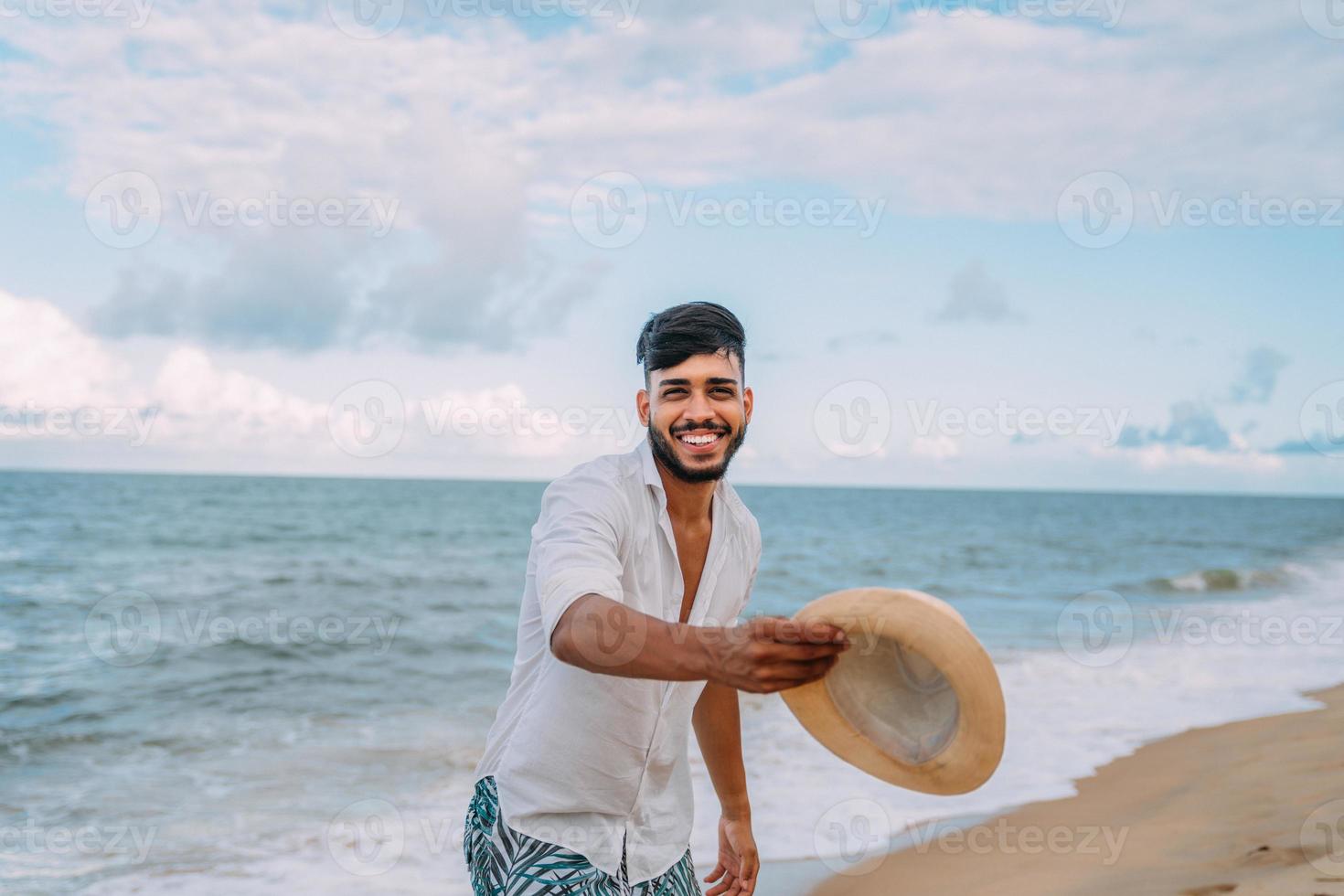 lateinamerikanischer mann lächelt und wirft seinen hut, der an einem schönen sommertag auf die kamera am strand schaut foto