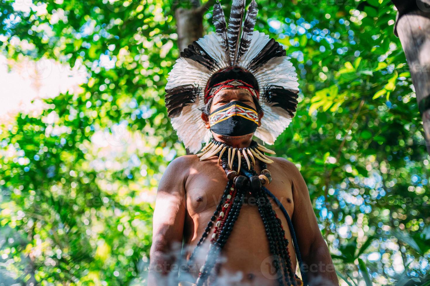 Schamane des Pataxo-Stammes. älterer inder mit federkopfschmuck und gesichtsmaske aufgrund der covid-19-pandemie. brasilianischer Indianer, der in die Kamera schaut foto