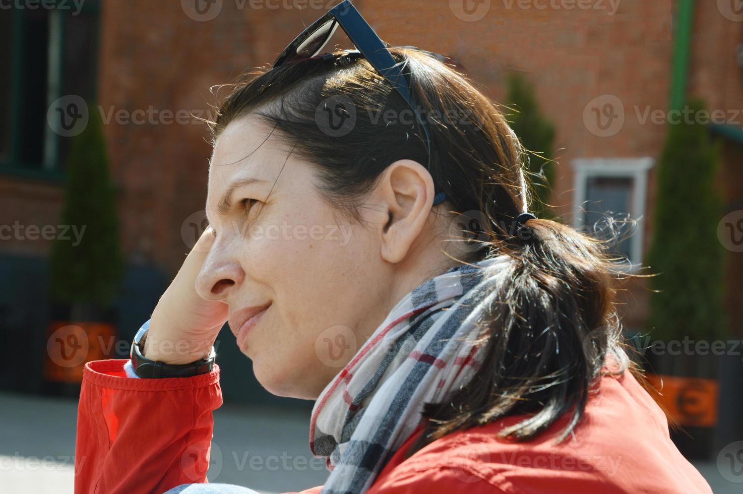 eine schöne frau mit einem nachdenklichen ausdruck ruht in der stadt. Nahaufnahmeporträt einer nachdenklichen Frau, die wegschaut und eine rote Jacke trägt, vor dem Hintergrund eines Stadtgebäudes. foto