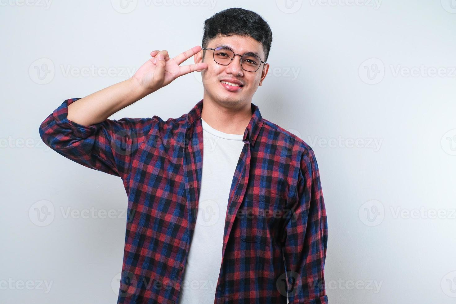 junger gutaussehender asiatischer kerl, der friedenssymbol mit den fingern über dem gesicht macht und fröhlich lächelt und den sieg zeigt foto