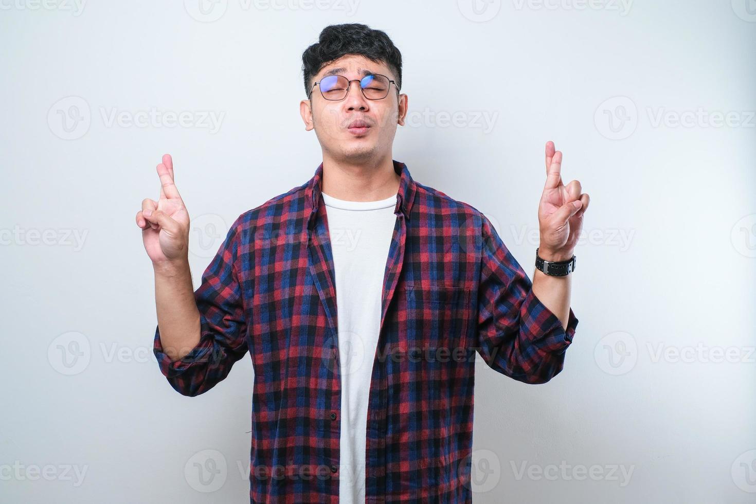 porträt eines jungen gutaussehenden asiatischen mannes mit lässigem hemd und brille, der die daumen drückt und für ein wunder wünscht und betet. foto