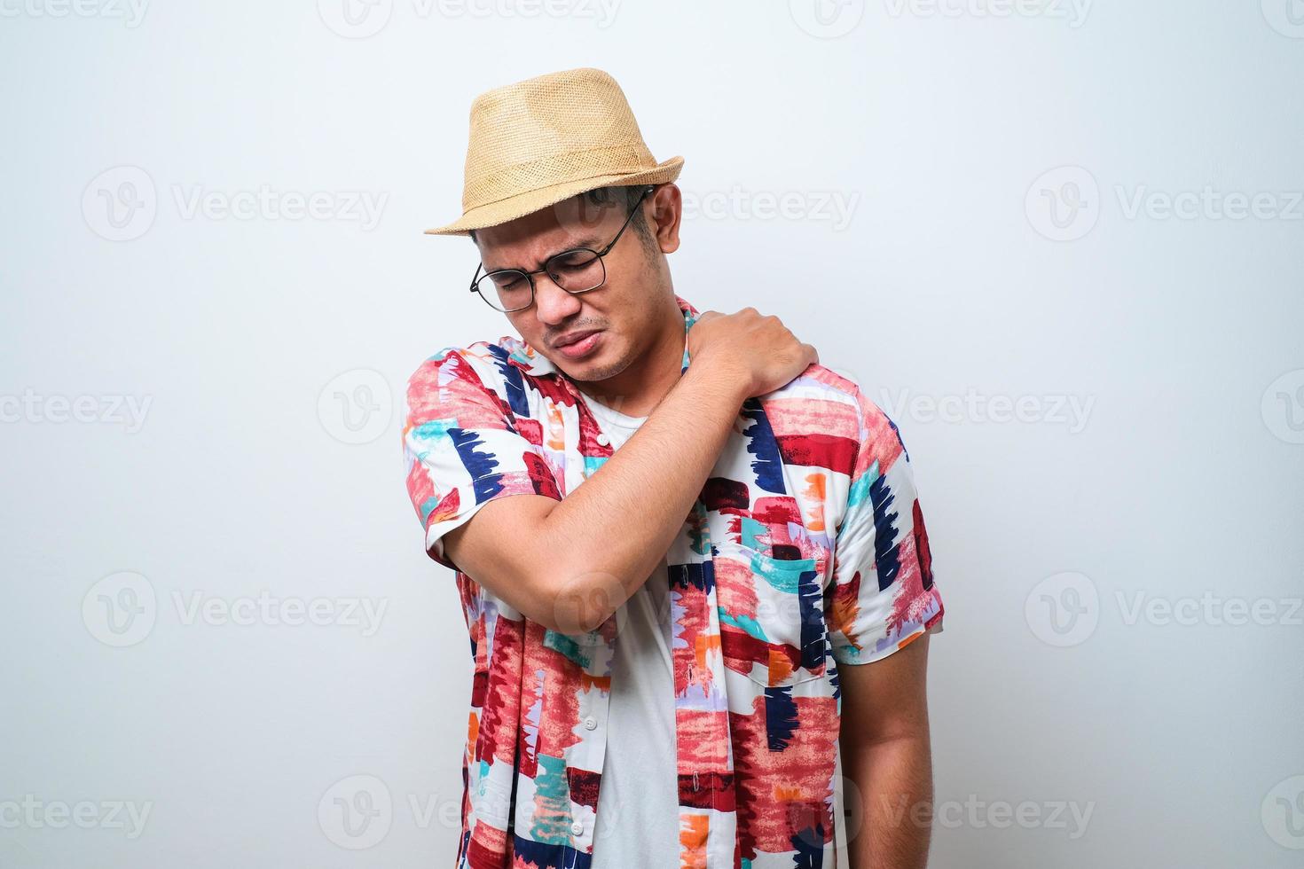 Der junge, gutaussehende asiatische Reisende, der ein lässiges Strandhemd und einen Eimerhut trägt, reibt sich die Schulter aufgrund der Rücken- und Schulterverletzung foto