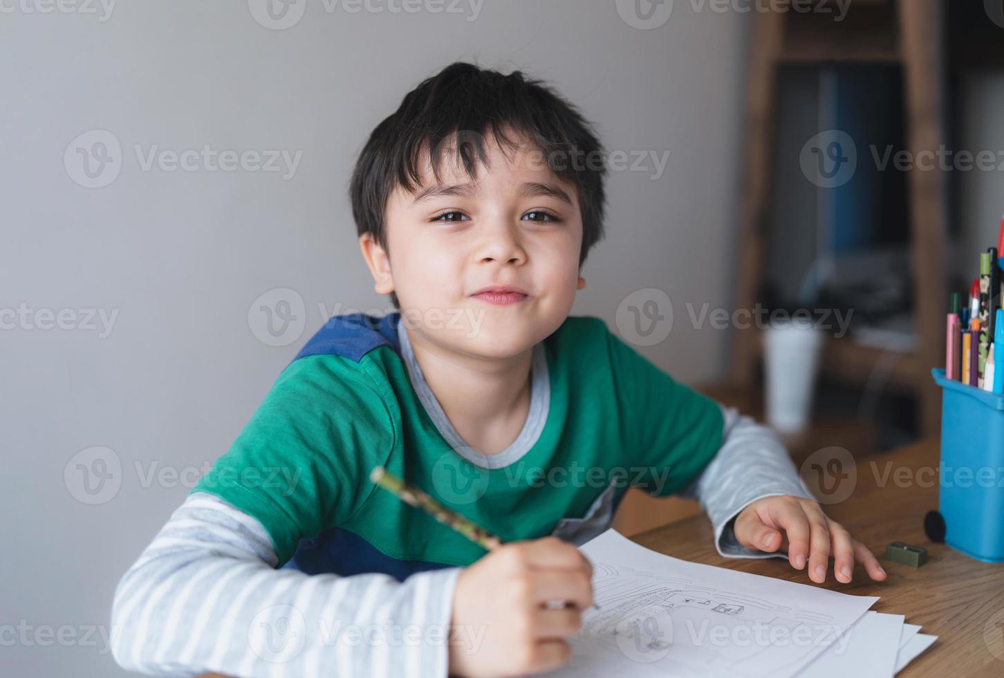 Fröhlicher Junge, der Bleistiftzeichnungen verwendet oder auf Papier skizziert, süßes Kind, das mit lächelndem Gesicht auf dem Tisch sitzt und Hausaufgaben macht, Kind genießt Kunst- und Handwerksaktivitäten zu Hause, Bildungskonzept foto