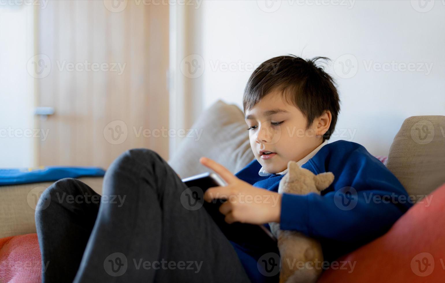 kleiner Junge, der ein Tablet-Spiel im Internet spielt, Kind, das auf dem  Sofa sitzt und online mit einem Freund spricht, Kind, das sich morgens im  Wohnzimmer entspannt, Kinder mit neuem Technologiekonzept 9711611