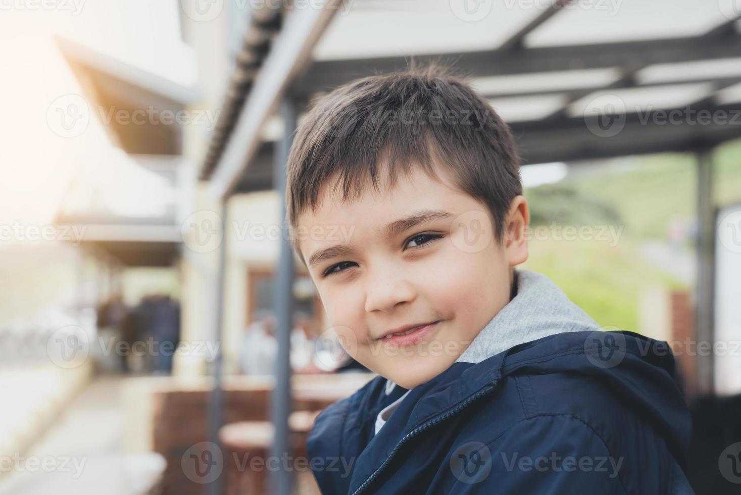 Kind sitzt an Bushaltestelle und wartet auf Schulbus. Porträt glücklicher kleiner Junge, der mit lächelndem Gesicht in die Kamera schaut, positives Kind, das an sonnigen Tagen im Frühling oder Sommer im Freien sitzt foto