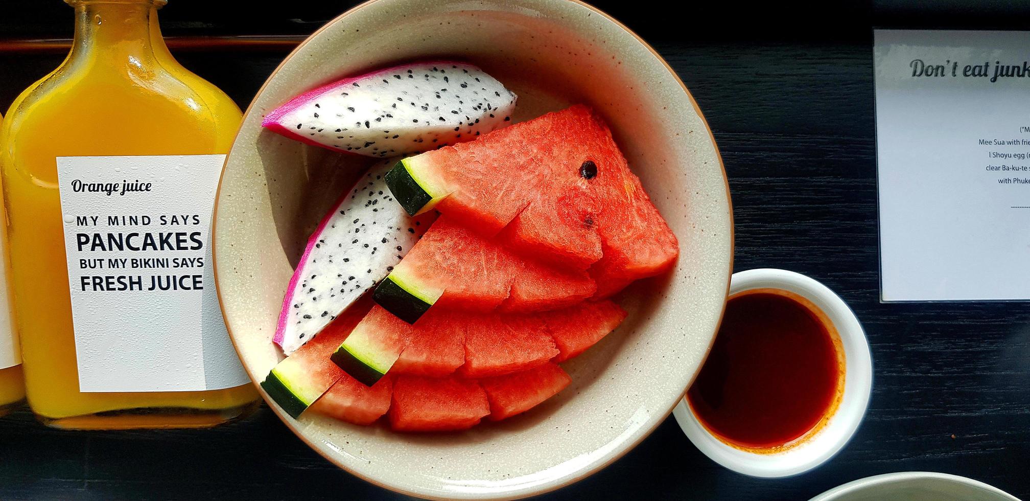 Draufsicht auf frisch geschnittene Wassermelone und Drachenfrucht in Teller mit Flasche Orangensaft auf schwarzem Holztisch. flache lage von frischem obst in der schale auf holzhintergrund. Reihe von gesunden Lebensmitteln und Ernährung foto