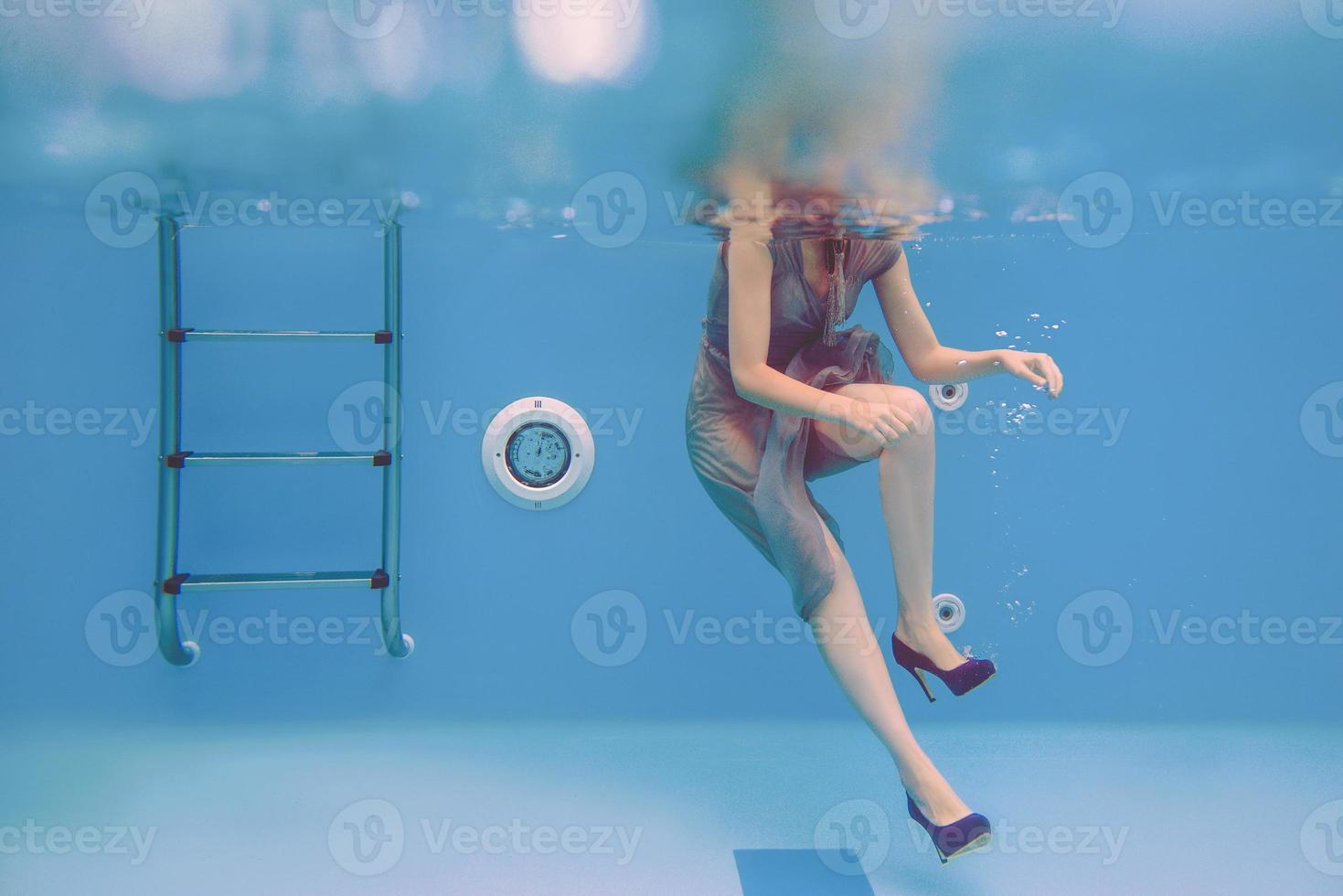 surreales kunstporträt einer jungen frau in grauem kleid, perlenschal, kleiner tasche, violetten high heels unter wasser im schwimmbad foto