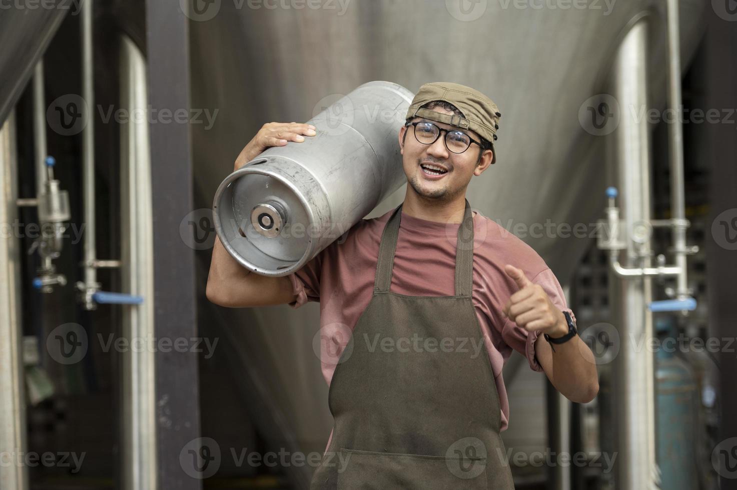 junger mann in lederschürze, der bierfass in der modernen brauerei hält, handwerksbrauereiarbeiter foto