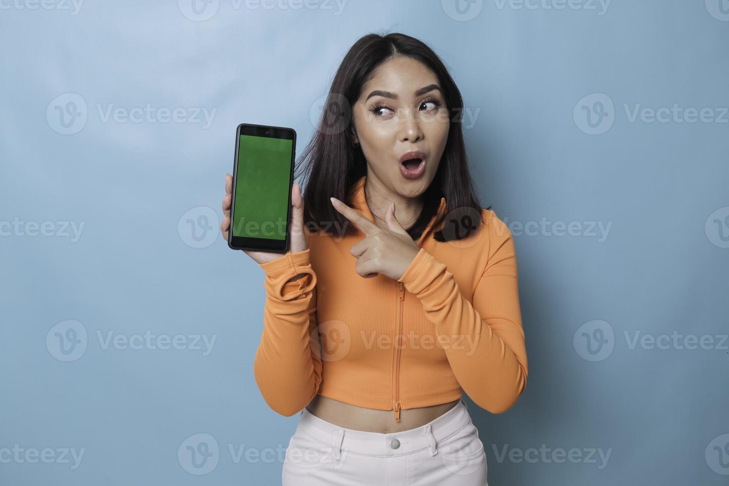 Eine überraschte junge Frau, die auf ihrem Smartphone in der Hand auf den Kopierbereich zeigt, isoliert auf blauem Hintergrund foto