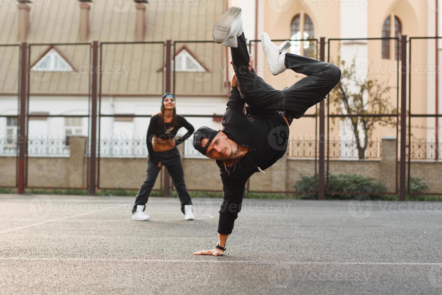 hübscher kerl in einer stilvollen schwarzen kleidung, die hip-hop tanzt. Tänzer, der auf seiner Hand steht. Das Mädchen ist hinter dem Kerl. foto