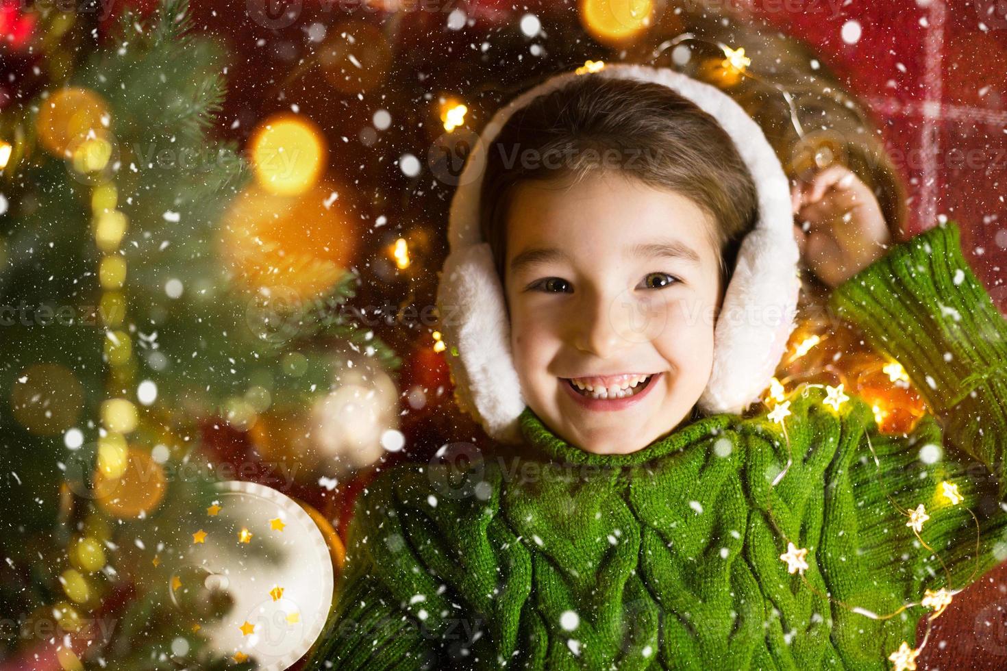 Ein fröhliches Mädchen mit Pelzkopfhörern liegt neben dem Weihnachtsbaum in Girlanden und Lichtern in einem warmen Pullover auf einer kuscheligen Decke. weihnachten und neujahr, feiertagsabend, magische stimmung foto