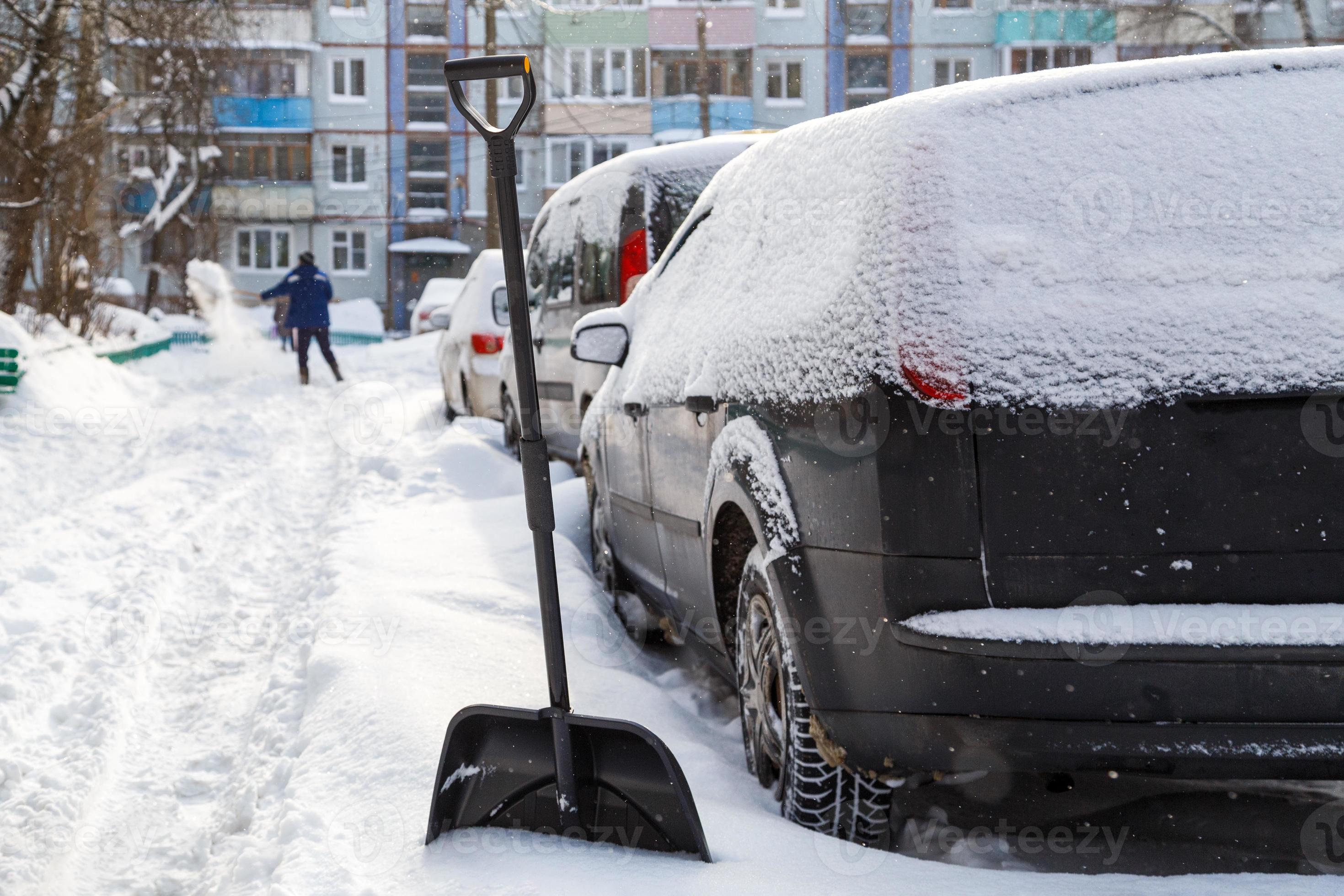 Schneeschaufel aus Kunststoff vor schneebedecktem Auto am sonnigen