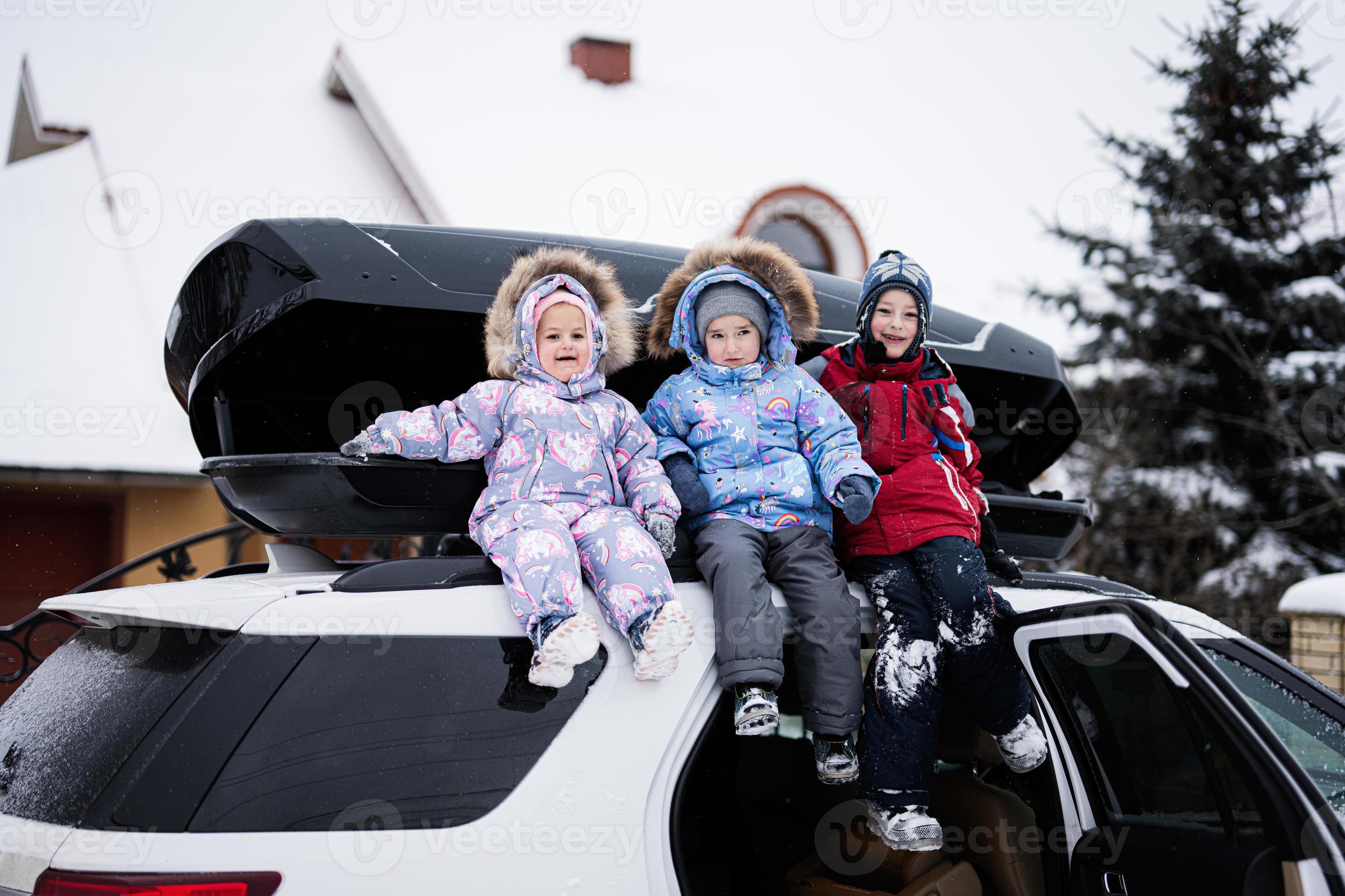 Drei Kinder sitzen im Winter auf einer Autodachbox gegen Haus