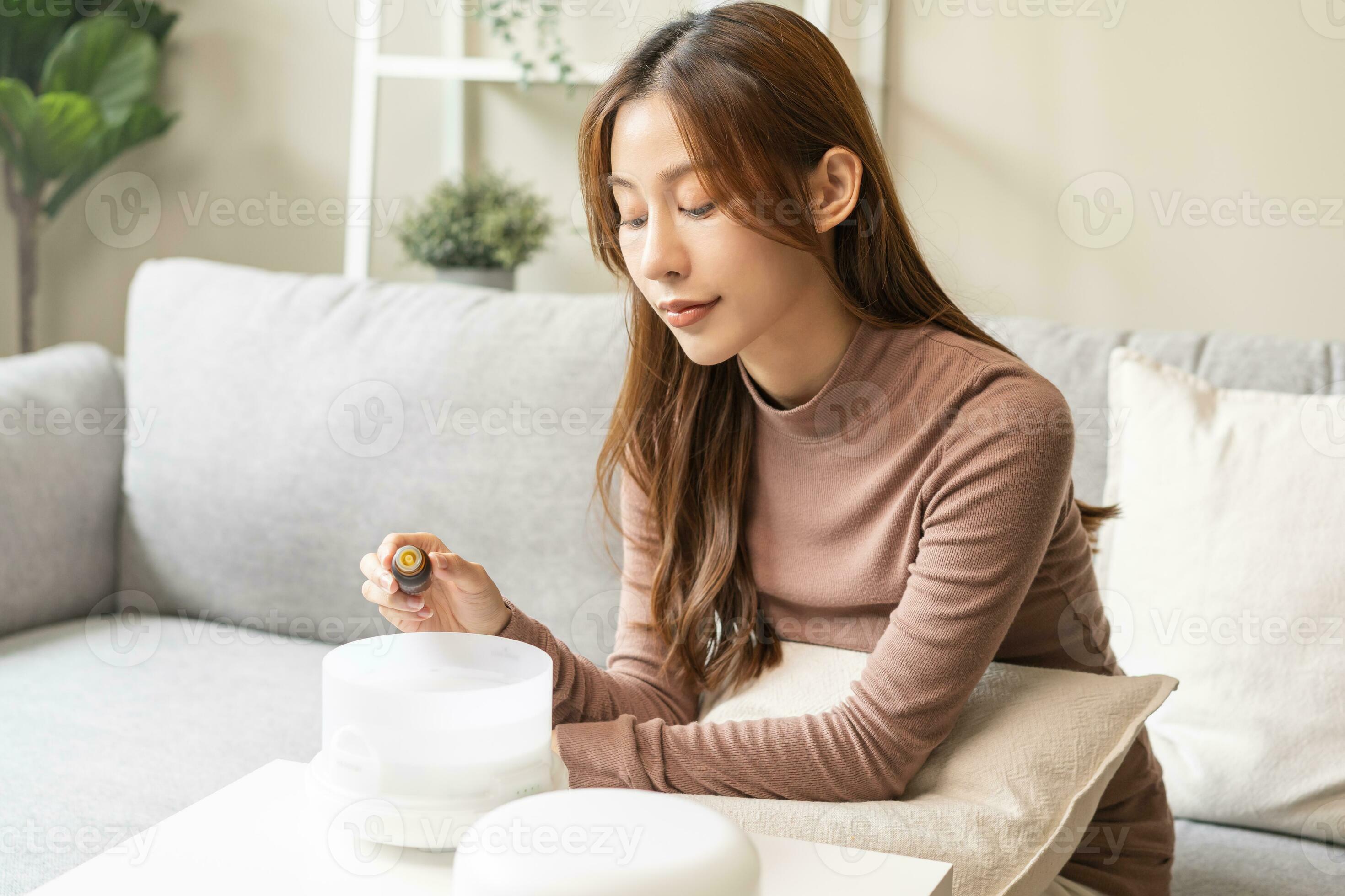 glücklich asiatisch jung Frau, Mädchen Hand halten Flasche von wesentlich  und fallen Öl in Luft Luftbefeuchter, genießen Aromatherapie Dampf Duft  Diffusor komfortabel während sich ausruhen im Leben Zimmer, Wohnung beim  heim. 25133872 Stock