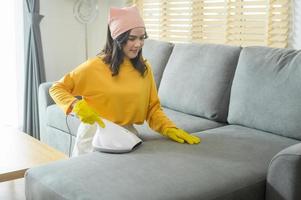 junge glückliche frau, die gelbe handschuhe trägt und ein sofa im wohnzimmer staubsaugt. foto