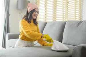 junge glückliche frau, die gelbe handschuhe trägt und ein sofa im wohnzimmer staubsaugt. foto