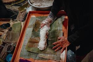 fokus alte frau mit traditionellem vietnamesischem kleid ao ba ba machen, verpacken tet kuchen, das vietnamesische mondneujahr tet essen im freien von händen. foto