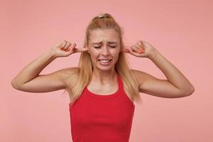 studiofoto einer gut aussehenden jungen frau mit lässiger frisur, die über rosa hintergrund steht, die ohren mit den händen bedeckt und faltet und versucht, laute geräusche zu vermeiden foto