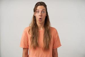 Innenfoto einer überraschten jungen Frau mit langen Haaren, die mit weit geöffneten Augen und gefalteten Lippen in die Kamera blickt und ein pfirsichfarbenes T-Shirt trägt, isoliert auf weißem Hintergrund foto