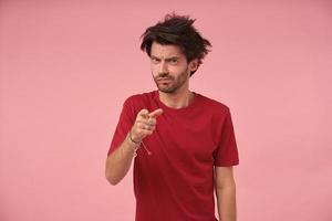 Innenaufnahme eines jungen gutaussehenden bärtigen Mannes mit wildem Haar, der mit dem Zeigefinger auf die Kamera zeigt, über rosa Hintergrund mit ernstem Gesicht steht und die Stirn runzelt und ein rotes T-Shirt trägt foto