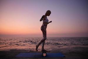 Outdoor-Foto von sportlich schlanken Frauen mit lässiger Frisur in sportlicher Kleidung, die auf einem Balance-Board stehen, bei Sonnenaufgang mit Balancer am Meer Sport treiben und die Hände heben, um das Gleichgewicht zu retten foto